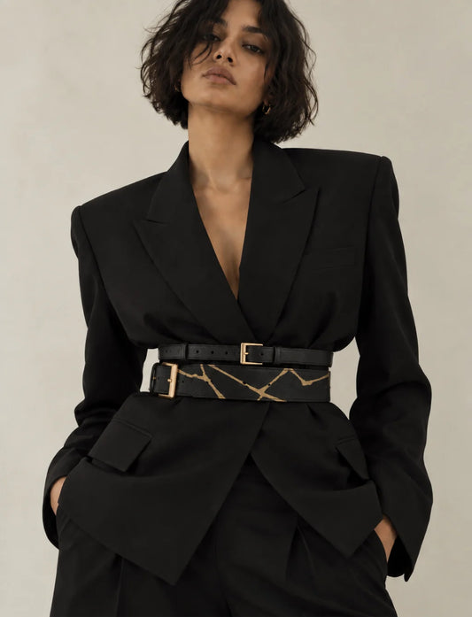 A model wearing a black structured blazer and matching trousers, styled with a wide black belt featuring a gold marble pattern and gold buckle, against a neutral beige background.